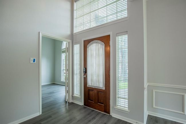 Bright entryway with a wooden front door featuring glass panels, flanked by tall windows. The area has light gray walls, elegant wainscoting, leading to an adjacent room with large windows. (home office on right side) | Image 9