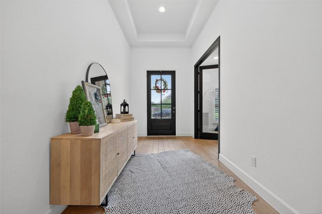 Welcoming front entry with a black framed glass door, tray ceiling, and wide hallway with wood-look tile floors. The neutral palette and clean finishes create a bright, modern feel upon entering the home. | Image 5