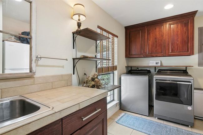 This photo showcases a laundry room with modern appliances, wooden cabinetry, and a tiled countertop with a sink. It features open shelving, a window for natural light, and a cozy rug on tiled flooring, creating a functional and inviting space. | Image 40
