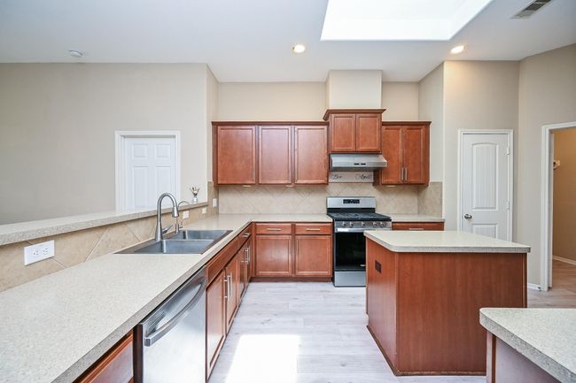 This kitchen features modern stainless steel appliances, ample cherry wood cabinetry, and a spacious island. The light countertops and tile backsplash enhance the airy feel, while a skylight adds natural brightness. | Image 20