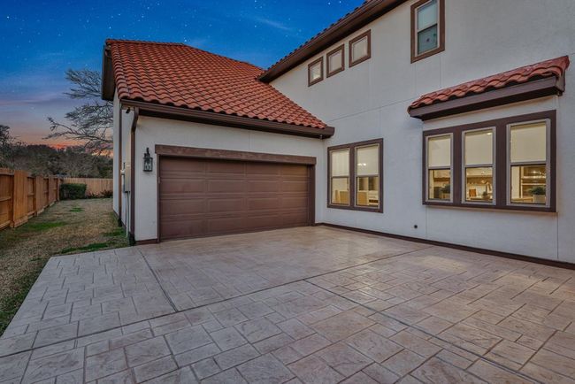 The modern home with a three-car tandem garage, featuring a tiled roof and a spacious paved driveway. The exterior has large, well-framed windows that hint at a bright interior, and the property is bordered by a wooden fence, offering privacy and a touch of nature. | Image 4