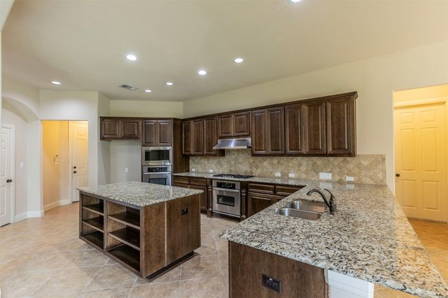 Kitchen with under cabinet range hood, stainless steel appliances, open shelves, backsplash, and dark brown cabinetry | Image 11