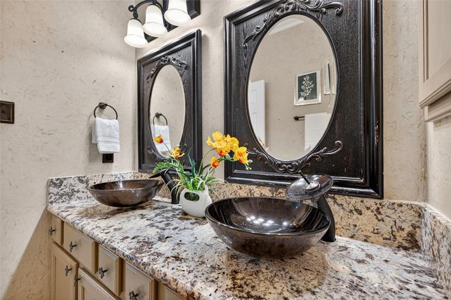 This bathroom features a granite countertop with dual vessel sinks, ornate black-framed mirrors, and elegant lighting. It’s accented with a neutral color scheme and decorative touches like a vase of flowers, offering a sophisticated and inviting space. | Image 23