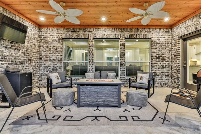 This inviting outdoor patio features a cozy seating area with a fire pit table, under a warm wood-paneled ceiling with ceiling fans. The brick walls add charm, while the mounted TV provides entertainment for gatherings. | Image 5