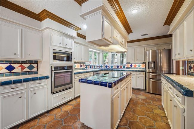 This kitchen features a spacious layout with white cabinetry, colorful tile countertops, and a central island with glass cooktop. Modern appliances, ample storage, and a stylish tiled backsplash, creates a bright and inviting space for cooking and entertaining. | Image 9