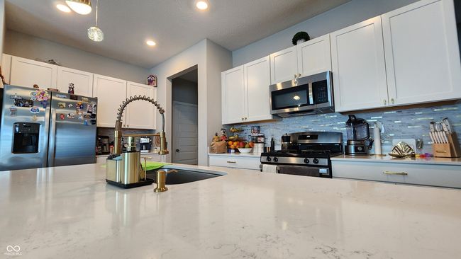 kitchen with stainless steel appliances, decorative backsplash, white cabinets, light stone counters, and hanging light fixtures | Image 6