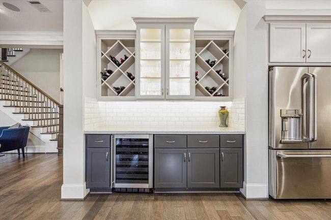 The bar has a wine refrigerator, a built-in wine rack, and additional cabinets for your storage needs. The light backsplash and under-cabinet lighting create a bright, inviting atmosphere, complemented by dark wood flooring. | Image 16