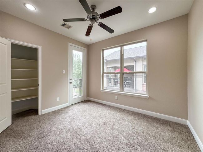 This secondary bedroom located on the second floor room features plush carpeting, a ceiling fan, and neutral walls. Natural light streams through a large window and a glass door, which opens to a small balcony. A closet with built-in shelves offers ample storage space. | Image 21