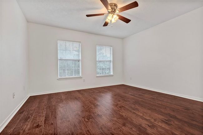 This room features clean white walls, rich wood flooring, and a ceiling fan with lights. Natural light fills the space through two windows with blinds, creating a bright and inviting atmosphere. | Image 28