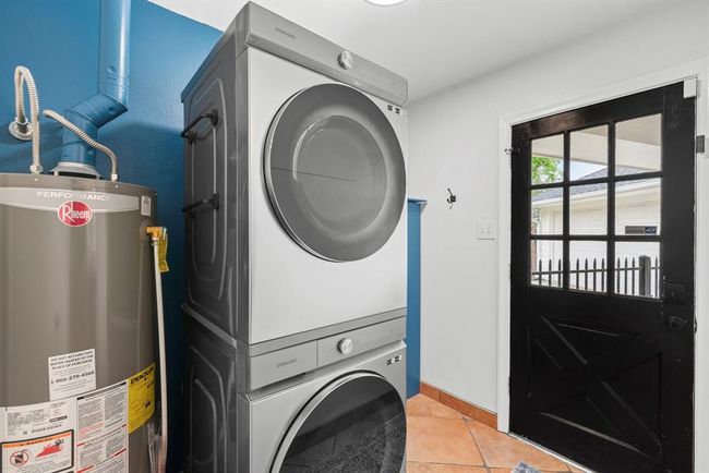 This photo shows a compact laundry room with a stacked washer and dryer next to the water heater. The space features a bright blue accent wall, terracotta tile flooring, and a black door with a window, leading outside. To the left of the water heater is the 1/2 bathroom. | Image 30