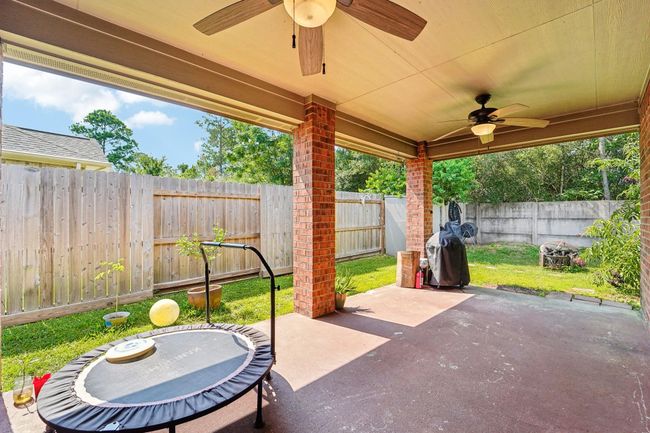 This photo showcases a covered patio with ceiling fans, brick pillars, and a view of a fenced backyard. The space includes a small trampoline and a grill, ideal for outdoor activities and relaxation. | Image 24