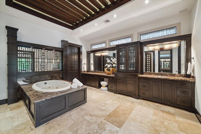 This luxurious primary bathroom offers a true spa-like experience, complete with a deep jetted tub set into a granite-surround platform and flanked by architectural wood columns and a mirrored wall with inlaid wood design. Dual vanities with built-in linen tower provide ample storage, while upper clerestory windows bathe the room in soft natural light. A dramatic coffered ceiling with lighted exposed beams adds architectural sophistication to this serene space. | Image 27