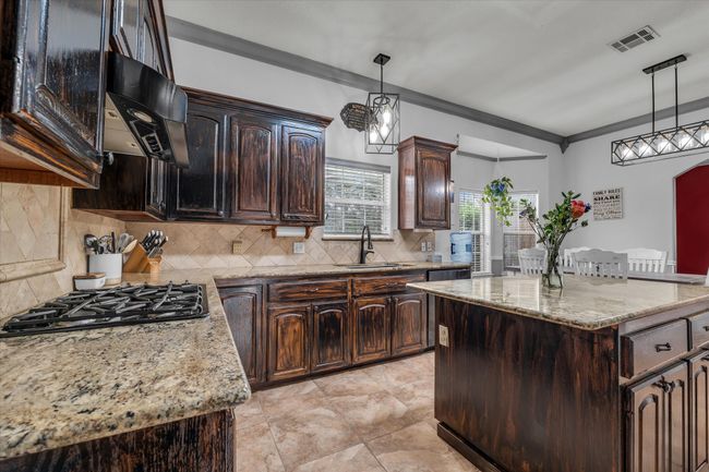 Kitchen with black gas stovetop, light stone counters, decorative light fixtures, dark brown cabinetry, and ornamental molding | Image 9