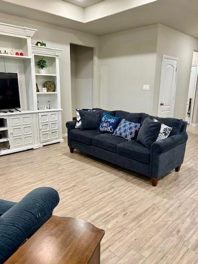 This cozy living room features wood-look tile flooring and a comfortable blue sofa with decorative pillows. It includes a white built-in entertainment center with shelving and a TV. The room has a neutral color palette and offers a welcoming space for relaxation. | Image 6