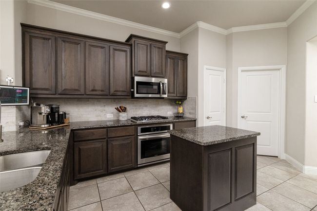 Kitchen featuring stainless steel appliances, dark brown cabinetry, dark stone counters, light backsplash, kitchen island, gas cooktop, electric oven, recessed lighting and pantry. | Image 7