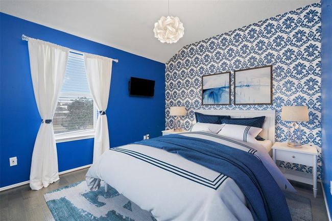 This bedroom features a bold blue and white color scheme with a patterned accent wall. It includes a cozy bed with matching linens, bedside tables with lamps, a wall-mounted TV, and a window with white curtains, creating a bright and inviting space. | Image 35
