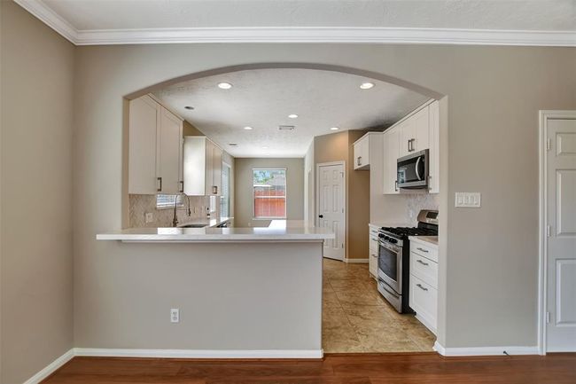 This kitchen features an open layout with a modern archway, sleek white cabinetry, and stainless steel appliances. The space is brightened by natural light from a large window, and the neutral color palette adds a warm, inviting feel. | Image 19