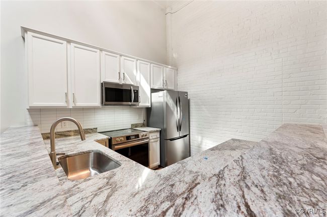 Kitchen with stainless steel appliances, light stone counters, decorative backsplash, white cabinets, and brick wall | Image 5
