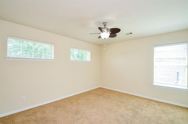 This secondary bedroom is spacious with carpeted flooring, neutral walls, and ample natural light from multiple windows. It features a ceiling fan and overhead lighting, making it bright and airy. | Image 25