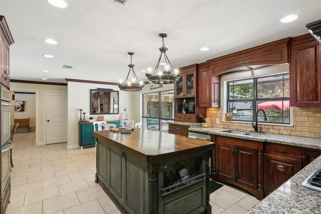This kitchen features rich wooden cabinetry, granite countertops, and a central island with a rustic finish. It includes modern appliances, a farmhouse sink, and a window overlooking the backyard. The open layout flows into a dining area, accentuated by elegant chandeliers and tiled flooring. | Image 22