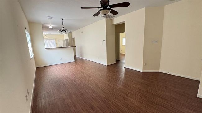 A spacious, open-concept living area with wood flooring, neutral walls, ceiling fans, and ample natural light, leading into a kitchen with a breakfast bar and pendant lighting. | Image 6