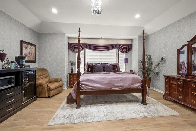 This is a spacious bedroom featuring a four-poster bed with elegant drapery, complemented by traditional wooden furniture and a cozy recliner. The room is well-lit and has a soothing color palette with patterned wallpaper, creating an inviting atmosphere. | Image 27