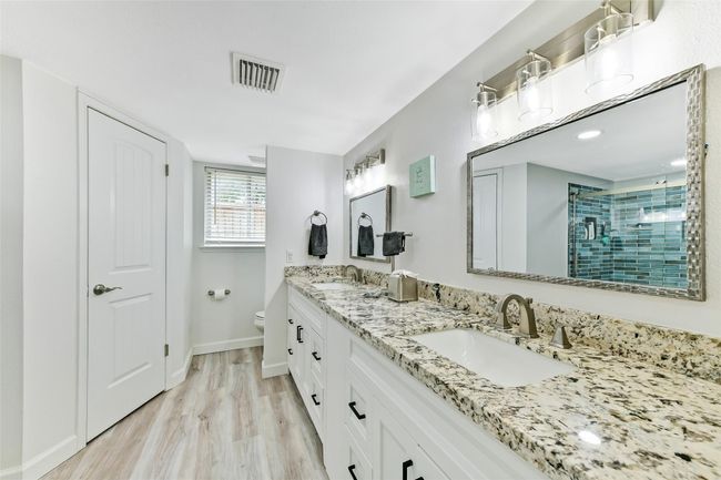 This bathroom features a double vanity with granite countertops, modern fixtures, and ample storage. It has a bright, airy feel with light wood flooring and a tiled walk-in shower. | Image 14