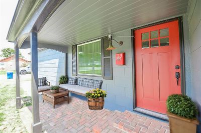 The home's entryway is inviting with a striking red door and matching shutters, set against freshly painted blue siding. This welcoming entrance is complemented by a traditional brick porch and a vintage-style outdoor lamp, enhancing the home's rustic charm and providing a warm welcome to all who visit. *Virtually staged.* | Image 3