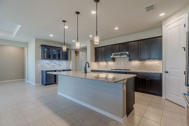 Kitchen with decorative backsplash, light stone counters, light tile patterned floors, a kitchen island with sink, and glass insert cabinets | Image 13