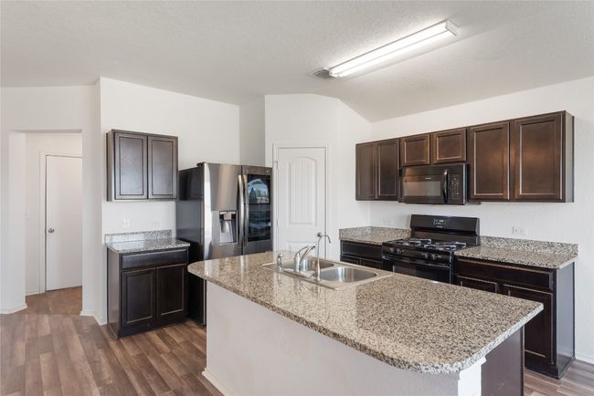 Kitchen with black appliances, dark brown cabinets, dark wood-type flooring, an island with sink, and light stone counters | Image 7