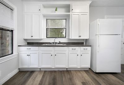 This is a clean, bright kitchen featuring white cabinetry, dark countertops, modern appliances, and wood-look flooring. Plenty of natural light comes in through the window over the sink. | Image 1