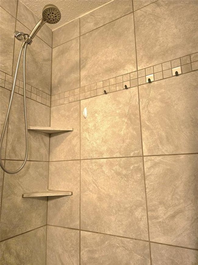 This photo shows a modern shower with large beige tiles, a handheld showerhead, and built-in corner shelves. The design is clean and minimalist, with a decorative tile border adding a touch of elegance. | Image 16