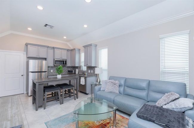 This photo highlights the stylish Next Gen suite, featuring a modern, open-concept living room and kitchen. The space includes sleek gray cabinetry, stainless steel appliances, and a cozy seating area with a light blue sofa adorned with decorative pillows. Large windows allow natural light to fill the room, creating a bright and welcoming atmosphere. Perfect for multi-generational living, this suite offers both comfort and convenience. | Image 30