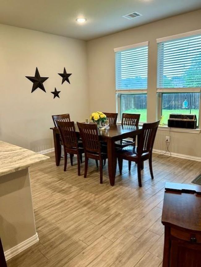 This photo showcases a cozy dining area with a wooden table and chairs, set against a backdrop of large windows offering ample natural light. The space features neutral walls adorned with decorative star accents and has a view of a green backyard. The flooring is a light wood-look tile, complementing the overall warm and inviting atmosphere. | Image 10