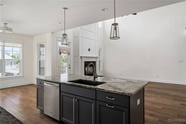 Kitchen featuring dark wood-type flooring, stainless steel dishwasher, light stone countertops, a center island with sink, and hanging light fixtures | Image 13