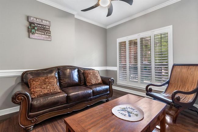 Cozy living room with elegant dark leather sofa, wooden chair, and coffee table. Features include crown molding, a ceiling fan, and large windows with plantation shutters for ample natural light. Neutral gray walls add a modern touch. | Image 5