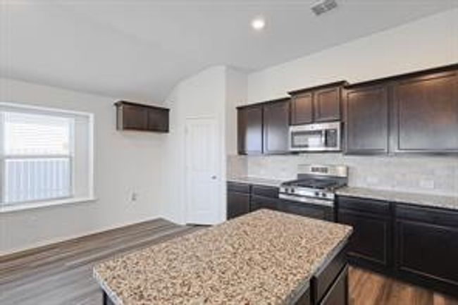 Kitchen featuring stainless steel appliances, a center island, wood finished floors, light stone countertops, and dark brown cabinetry | Image 6