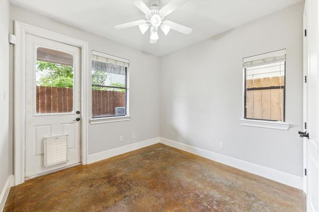 Bright room with concrete floors, neutral walls, and a ceiling fan. It features a door with a pet flap for outdoor access to the turfed backyard. | Image 23