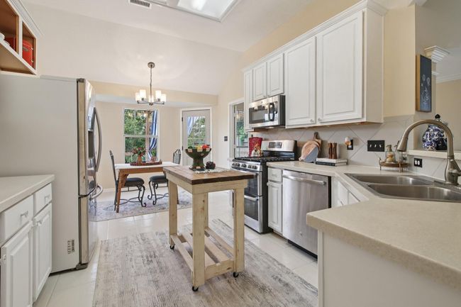 This photo showcases a bright, modern kitchen with white cabinetry, and stainless steel appliances. It opens to a cozy dining area with large windows, offering a pleasant view of the outdoors. | Image 13