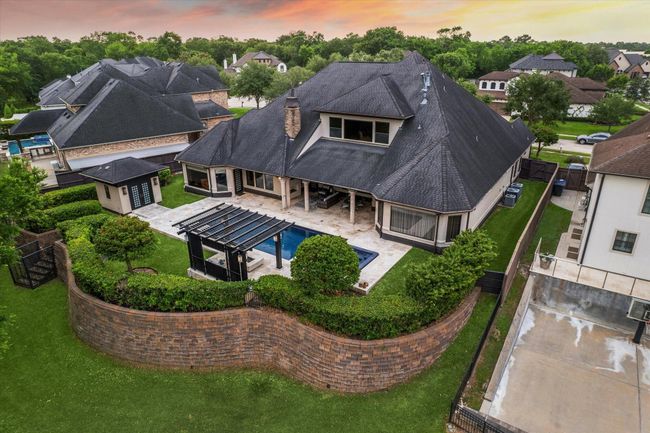 This elevated view captures the full scope of the home's resort-style backyard, artfully enclosed by a curved retaining wall that enhances both privacy and visual appeal. The seamless connection between the main residence, Zen-style pool house, and outdoor entertaining spaces reflects thoughtful design. Manicured hedges, a pergola-covered fire pit lounge, and the sleek pool and spa create a luxurious outdoor living experience in perfect harmony with the natural surroundings. | Image 49