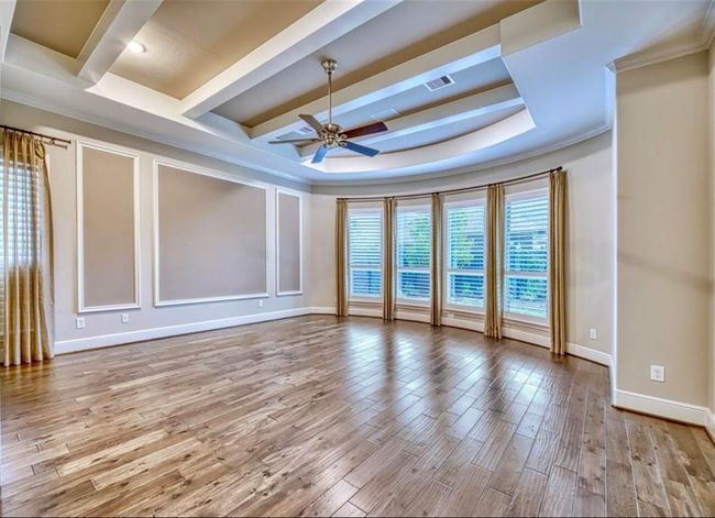 This spacious room features elegant tray ceilings with recessed lighting, a stylish ceiling fan, and large bay windows with plantation shutters. The hardwood floors add warmth, and the neutral wall colors provide a versatile backdrop for any decor. | Image 27