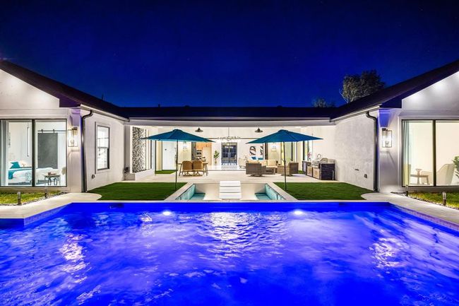 A stunning view of this contemporary home's backyard oasis showcases a beautifully illuminated pool leading up to a covered patio with built-in lighting and ceiling fans. The outdoor area includes a sunken fire pit lounge, lush grass, and a fully equipped outdoor kitchen—perfectly designed for seamless indoor-outdoor living and upscale entertaining under the stars. | Image 17