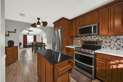 This is a spacious kitchen with rich wooden cabinetry, modern stainless-steel appliances, a central island, and a tile backsplash. The flooring is wood-look tile, and there's an arched opening leading to the dining area with ample natural light. | Image 3