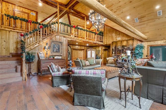 This cozy living area features a warm, rustic design with wood-paneled walls and ceilings. The space includes a grand chandelier, comfortable seating, and decorative touches like garland and artwork. A staircase leads to a loft area, adding charm and functionality to the space. | Image 24