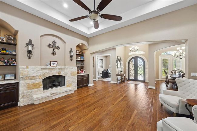 This inviting living area features a cozy stone fireplace, elegant ceiling fan, and warm wood flooring. Decorative arches and built-in shelves add charm, while large doors and windows provide ample natural light. The open layout flows seamlessly into an adjacent dining area. | Image 5
