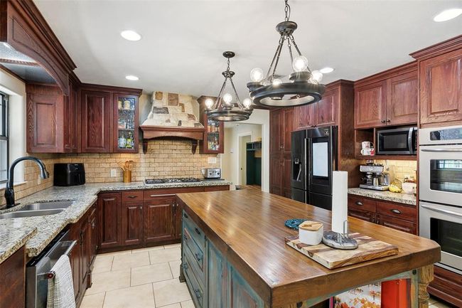 This kitchen features rich wooden cabinetry and a large central island with a wooden countertop. It boasts modern appliances, including a double oven and a sleek black refrigerator. The stone backsplash and decorative range hood add a rustic touch, while ample lighting and granite counters enhance functionality and style. | Image 20