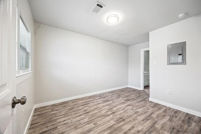 This photo shows a small, bright room with light-colored walls and wood-style flooring. It features a window for natural light, a ceiling light fixture, and an electrical panel on one wall. There's a doorway leading to another room. | Image 12