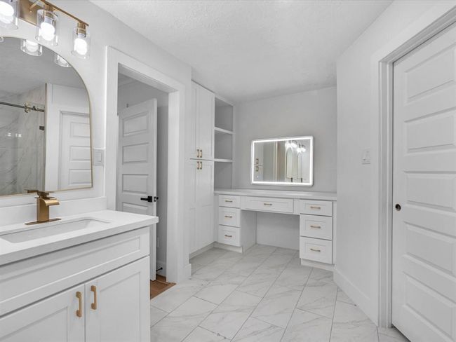 This bathroom features a modern design with white cabinetry, gold hardware, and a sleek marble-like tile floor. It includes a spacious vanity with a large illuminated mirror, ample storage, and elegant lighting fixtures for a bright and airy feel. | Image 24