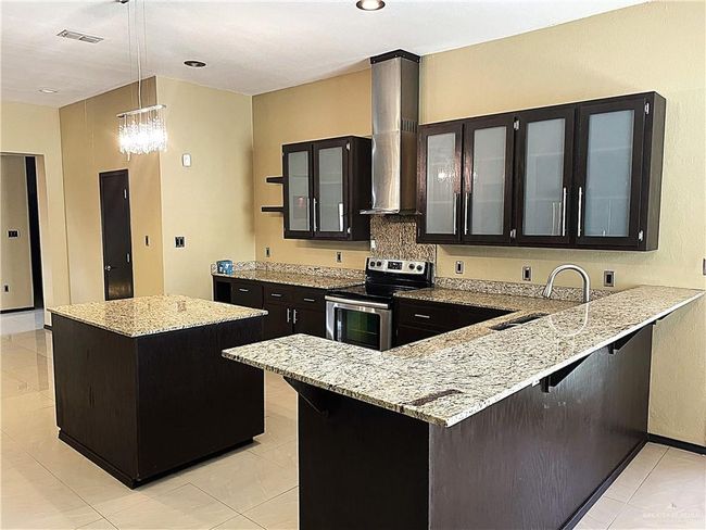 Kitchen featuring stainless steel electric stove, wall chimney exhaust hood, a sink, dark cabinets, and a kitchen island | Image 6