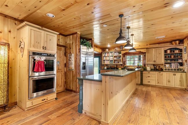 This spacious kitchen features rustic wood paneling, modern stainless steel appliances, and a large island with granite countertops. It has ample storage, warm lighting, and a cozy, welcoming atmosphere. | Image 16